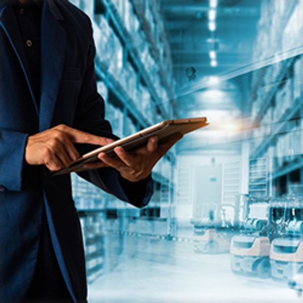 A closeup of a man's hand scrolling on a digital tablet with a warehouse in the background. 