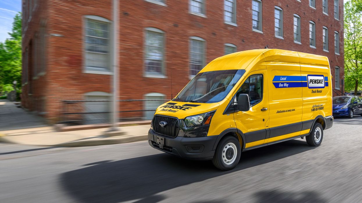 Yellow cargo van with Penske branding driving down street passing a brick building